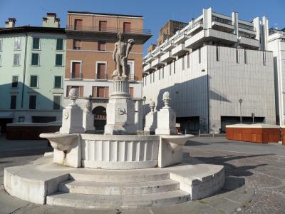 A stone fountain featuring a statue of a figure holding a cornucopia, situated in a paved public square with multi-story buildings in the background.