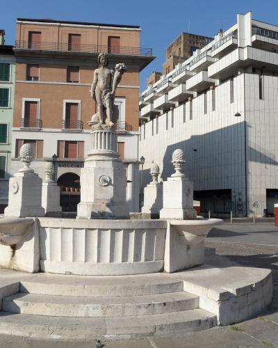 A stone fountain featuring a statue of a figure holding a cornucopia, situated in a paved public square with multi-story buildings in the background.