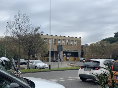 A street-level view of a large, two-story brick building with the words "MERCATO ORTOFRUTTICOLO" visible on its facade, situated in an urban area with cars, motorcycles, and trees under an overcast sky.