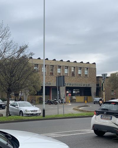 A street-level view of a large, two-story brick building with the words "MERCATO ORTOFRUTTICOLO" visible on its facade, situated in an urban area with cars, motorcycles, and trees under an overcast sky.