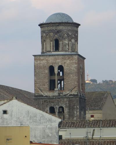 A tall, historic brick bell tower (campanile) with arched openings and a domed top, set against a cloudy sky with surrounding rooftops and distant hills.