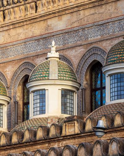 A close-up architectural detail showing three small domed lanterns with green and yellow glazed tiles, set against a stone facade featuring Moorish-style horseshoe arches and intricate geometric tilework.