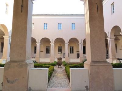 The image shows a sunlit courtyard with a gravel path, manicured hedges, and a central stone basin, framed by large square pillars and an arcade of arches leading to a pale pink building facade.