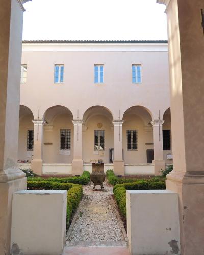 The image shows a sunlit courtyard with a gravel path, manicured hedges, and a central stone basin, framed by large square pillars and an arcade of arches leading to a pale pink building facade.