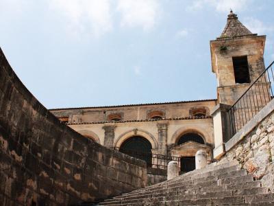 A low-angle view of a Baroque church facade with a bell tower, accessed by a wide stone staircase flanked by high retaining walls under a blue sky.