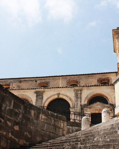 A low-angle view of a Baroque church facade with a bell tower, accessed by a wide stone staircase flanked by high retaining walls under a blue sky.