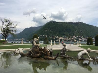 A fountain featuring classical statues of figures and sea creatures in a park, with a lake and a large green hill in the background.