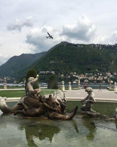 A fountain featuring classical statues of figures and sea creatures in a park, with a lake and a large green hill in the background.