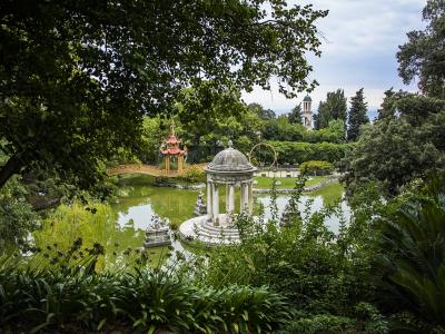 A view of the gardens of Villa Durazzo Pallavicini in Genoa, featuring a central white pavilion on an island in a pond, a distinctive red and gold Chinese-style pagoda bridge, and a church tower in the background.