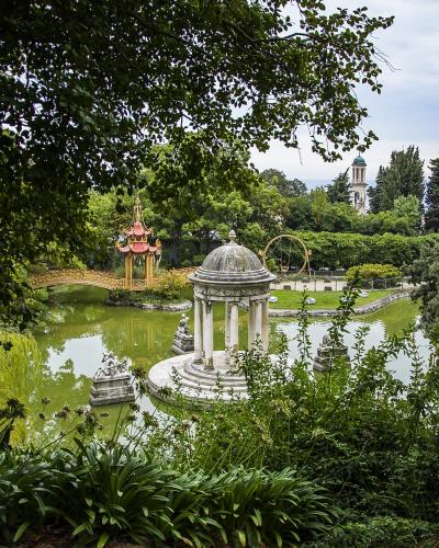 A view of the gardens of Villa Durazzo Pallavicini in Genoa, featuring a central white pavilion on an island in a pond, a distinctive red and gold Chinese-style pagoda bridge, and a church tower in the background.