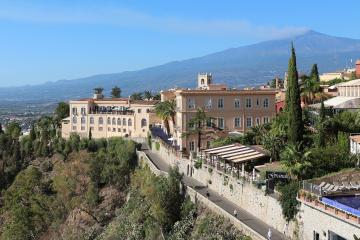A large, multi-story hotel complex with terracotta-colored facades and arched windows is situated on a hillside overlooking a coastal plain, with a prominent mountain (Mount Etna) in the background; signage for "Grand Hotel" is visible near a pool