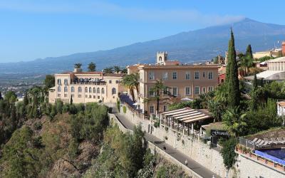 A large, multi-story hotel complex with terracotta-colored facades and arched windows is situated on a hillside overlooking a coastal plain, with a prominent mountain (Mount Etna) in the background; signage for "Grand Hotel" is visible near a pool
