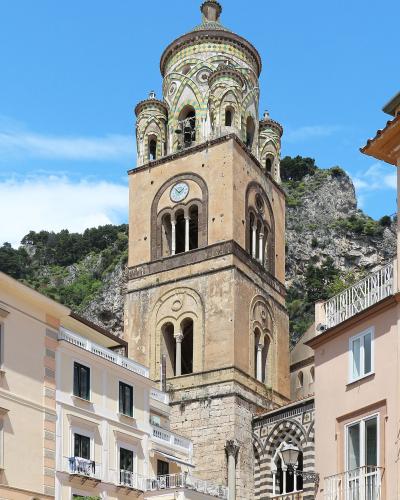 The image shows the bell tower (campanile) of the Cathedral of Saint Andrew (Duomo di Sant'Andrea) in Amalfi, Italy, featuring its distinctive Byzantine-style mosaic dome and clock face, set against a backdrop of steep, green-covered cliffs and ad