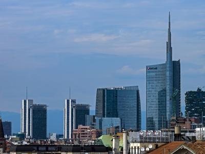 A cityscape showing the Unicredit Tower and the Bosco Verticale towers in Milan's Porta Nuova district, with other modern skyscrapers and foreground rooftops under a blue sky.