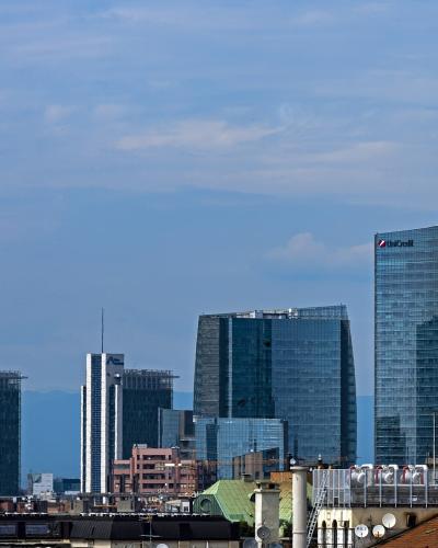 A cityscape showing the Unicredit Tower and the Bosco Verticale towers in Milan's Porta Nuova district, with other modern skyscrapers and foreground rooftops under a blue sky.
