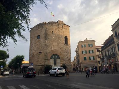 A large, cylindrical stone tower with an arched gateway at its base, an Italian flag flying from the top, and modern vehicles and pedestrians in a paved plaza setting.