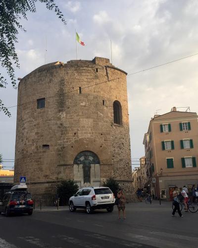 A large, cylindrical stone tower with an arched gateway at its base, an Italian flag flying from the top, and modern vehicles and pedestrians in a paved plaza setting.
