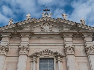 The image shows the upper facade of a large, light-colored stone cathedral with a triangular pediment, a central cross, and multiple white statues of saints positioned along the roofline and corners, set against a blue sky with clouds.