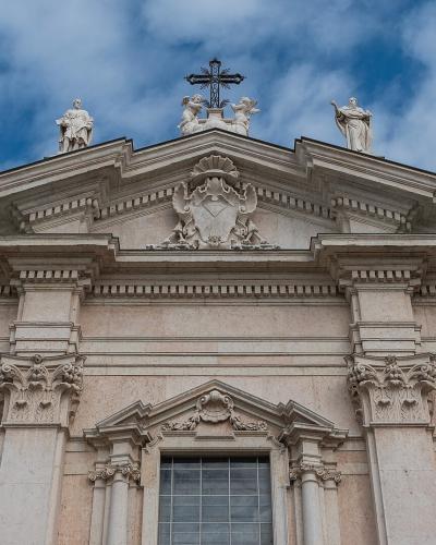 The image shows the upper facade of a large, light-colored stone cathedral with a triangular pediment, a central cross, and multiple white statues of saints positioned along the roofline and corners, set against a blue sky with clouds.
