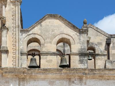 The image shows the upper facade and bell gables of a historic stone church with three arched openings containing bells, set against a blue sky.