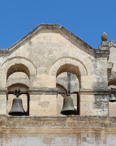 The image shows the upper facade and bell gables of a historic stone church with three arched openings containing bells, set against a blue sky.
