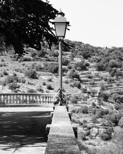 A black-and-white photograph showing a stone terrace with a balustrade, a vintage-style lamppost, a bench under a large tree, and a view of a terraced, hilly landscape in the background.