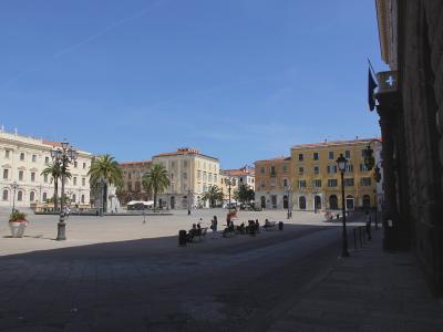 A wide, sunlit civic square paved with stone, featuring classical European-style buildings with arched ground floors and shuttered windows, palm trees, ornate lampposts, benches with people sitting, and a large clock tower building on the left; no
