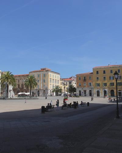 A wide, sunlit civic square paved with stone, featuring classical European-style buildings with arched ground floors and shuttered windows, palm trees, ornate lampposts, benches with people sitting, and a large clock tower building on the left; no