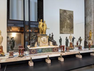 A museum gallery interior featuring a long display table with numerous bronze and gilded sculptures, including a central golden statue group on a marble pedestal, with a large painting on the wall and a glass elevator structure to the left.