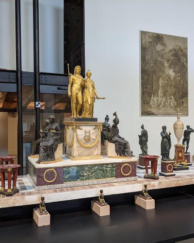 A museum gallery interior featuring a long display table with numerous bronze and gilded sculptures, including a central golden statue group on a marble pedestal, with a large painting on the wall and a glass elevator structure to the left.