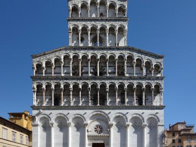 A full-frontal view of the ornate Romanesque facade of a white marble church with multiple tiers of blind arcades, topped by a statue of the Archangel Michael, situated in a paved public square under a clear blue sky.
