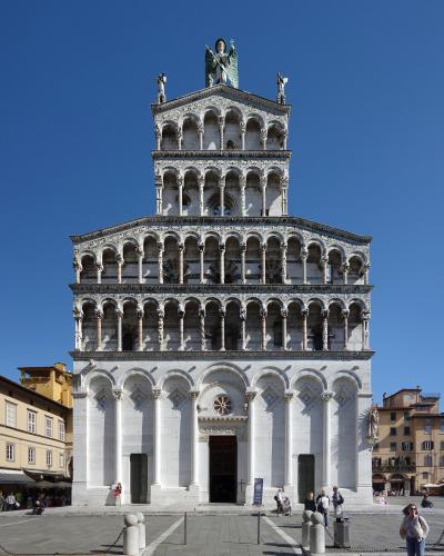 A full-frontal view of the ornate Romanesque facade of a white marble church with multiple tiers of blind arcades, topped by a statue of the Archangel Michael, situated in a paved public square under a clear blue sky.