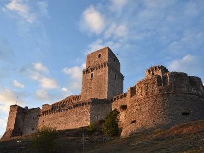 A low-angle photograph of a large medieval stone fortress with a prominent square tower and crenellated walls, situated on a grassy hill under a blue sky with scattered clouds.
