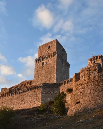 A low-angle photograph of a large medieval stone fortress with a prominent square tower and crenellated walls, situated on a grassy hill under a blue sky with scattered clouds.