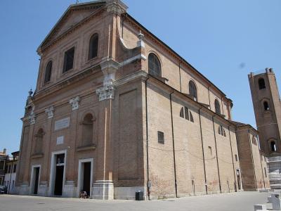A low-angle exterior shot of a large, brick-faced Renaissance-style church with a prominent triangular pediment, arched windows, and a tall, square brick bell tower to the right, set against a clear blue sky in an open paved square.