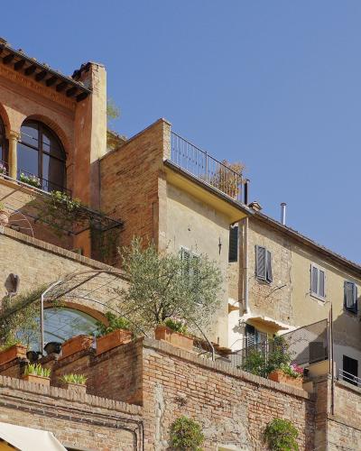 A sunlit street scene in a historic Italian hill town, featuring terracotta-roofed buildings with arched windows, green shutters, brick retaining walls, potted plants, and a clear blue sky; no visible signage or text is present.