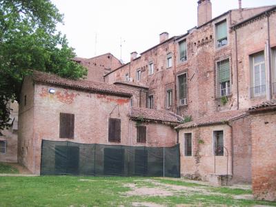 A grassy courtyard surrounded by aged red-brick buildings with shuttered windows and a black mesh fence in the foreground, likely part of a historic palace complex in Italy.
