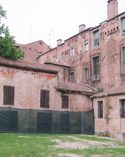 A grassy courtyard surrounded by aged red-brick buildings with shuttered windows and a black mesh fence in the foreground, likely part of a historic palace complex in Italy.