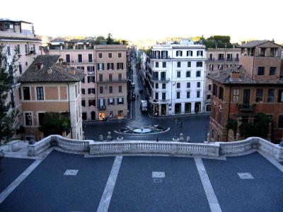 A high-angle view looking down the Spanish Steps in Rome towards the Piazza di Spagna and the Fontana della Barcaccia, with Via dei Conciatori visible as the street extending straight ahead from the square.