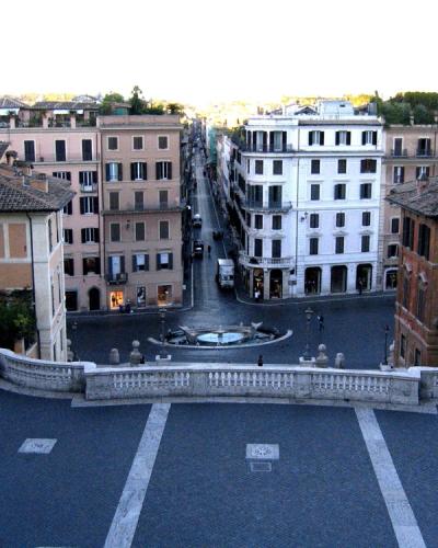 A high-angle view looking down the Spanish Steps in Rome towards the Piazza di Spagna and the Fontana della Barcaccia, with Via dei Conciatori visible as the street extending straight ahead from the square.
