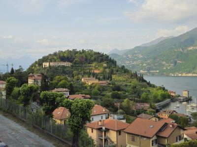 A scenic elevated view of a lakeside town with terracotta-roofed buildings, lush green hills, a large villa on a hilltop, and a small harbor with boats; no visible signage or text identifying the location.
