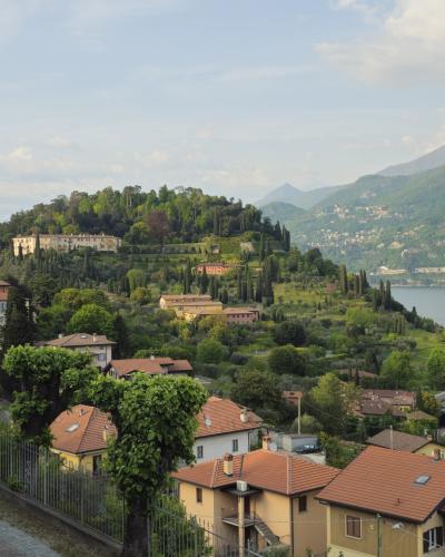 A scenic elevated view of a lakeside town with terracotta-roofed buildings, lush green hills, a large villa on a hilltop, and a small harbor with boats; no visible signage or text identifying the location.
