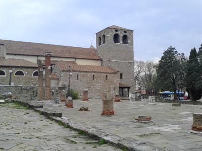 The image shows the ancient Roman forum ruins in Cividale del Friuli, Italy, featuring stone column bases and a prominent medieval bell tower (likely part of the Cathedral of Santa Maria Assunta) in the background.
