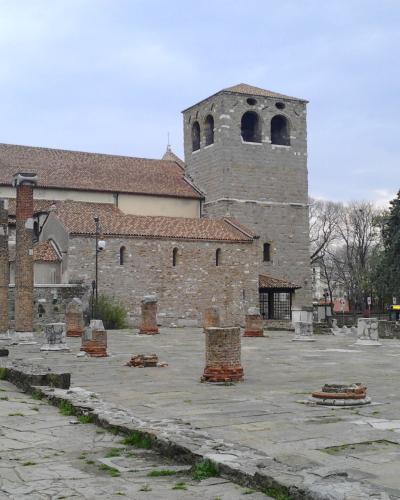The image shows the ancient Roman forum ruins in Cividale del Friuli, Italy, featuring stone column bases and a prominent medieval bell tower (likely part of the Cathedral of Santa Maria Assunta) in the background.