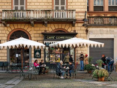 The image shows the exterior of a historic café with outdoor seating under white umbrellas, featuring a black sign reading "CAFFÈ CONFETTERIA" and a chalkboard menu advertising "Bicerin," set against a yellow stucco building with arched windows an