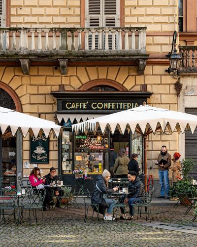 The image shows the exterior of a historic café with outdoor seating under white umbrellas, featuring a black sign reading "CAFFÈ CONFETTERIA" and a chalkboard menu advertising "Bicerin," set against a yellow stucco building with arched windows an