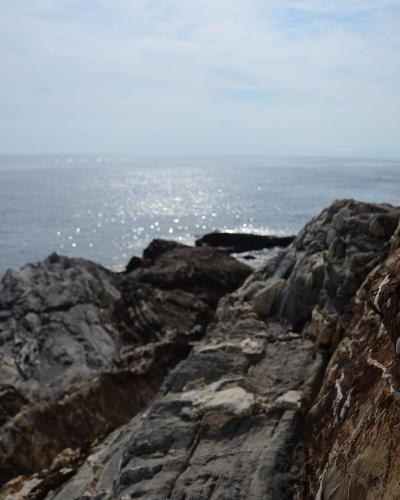 A close-up view of rugged, stratified rock formations along a coastline with the sunlit sea in the background.