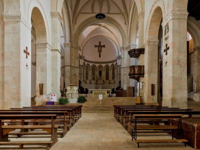 The interior of a stone church with arched columns, wooden pews, a central aisle, a suspended crucifix, and an altar area at the far end.