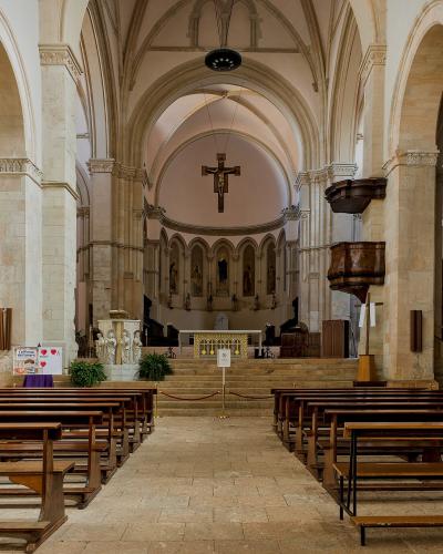 The interior of a stone church with arched columns, wooden pews, a central aisle, a suspended crucifix, and an altar area at the far end.