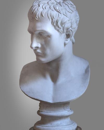 A white marble bust of a man with short, wavy hair shown in profile on a decorative pedestal against a plain grey background.